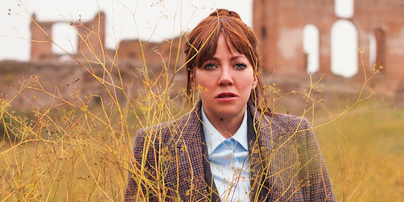 Diane Morgan as Philomena Cunk standing behind foliage and in front of some ruins