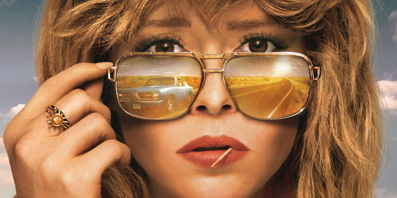 close-up of Natasha Lyonne lowering her sunglasses, a vintage blue car and open road are reflected in the lenses