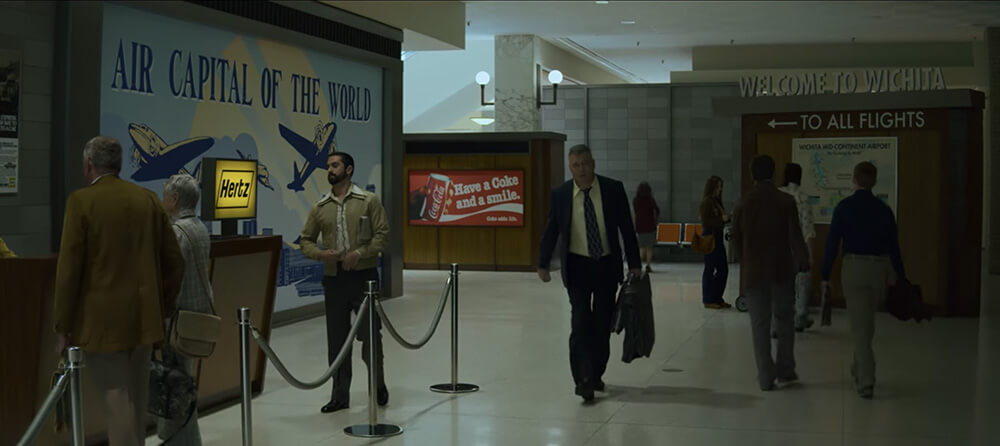 Tench walks through the airport and passes a Coca-Cola advertisement that says “Have a COke and a smile”
