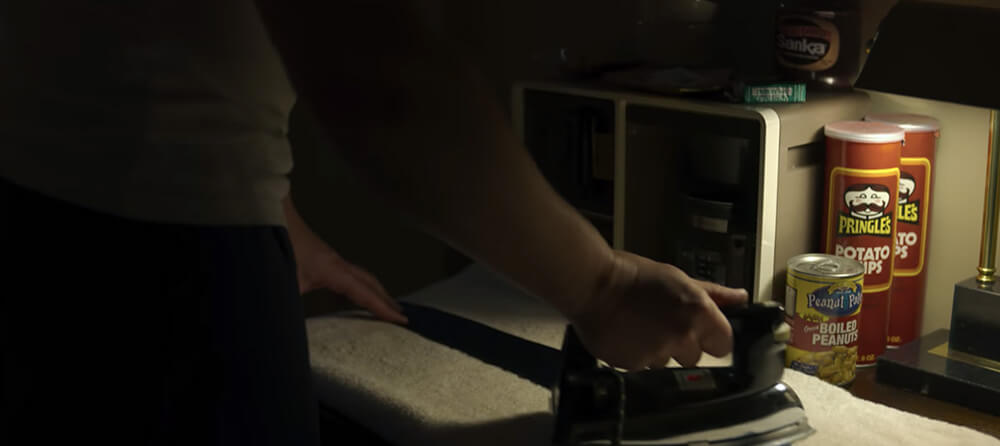 a hand irons a shirt in the dark and a nearby lamp illuminates a can of boiled peanuts and two Pringles containers