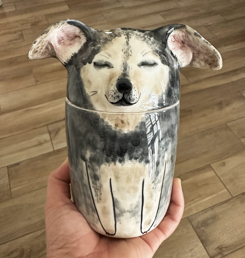 a ceramic cylindrical urn with a rounded top that looks like a little brown and black dog