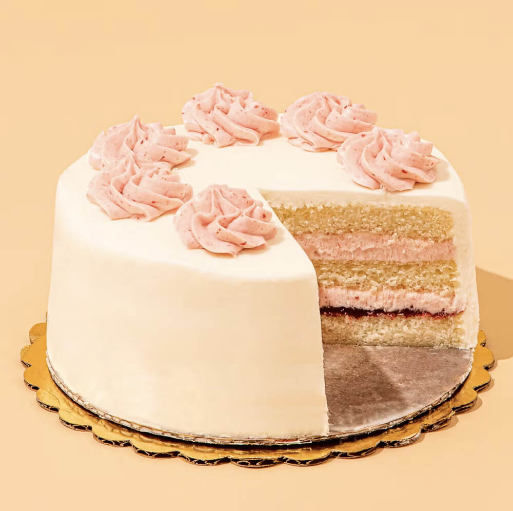 round white cake with pink frosted decoration, a slice is cut out to show layers of white cake, pink frosting, and raspberry jam