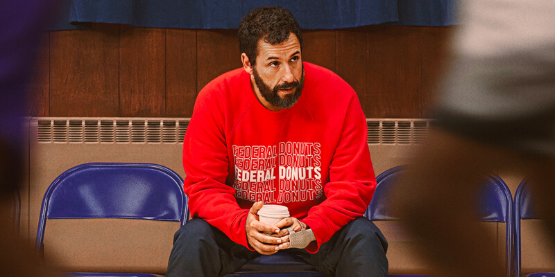 Adam Sandler holding a coffee on the sidelines of a basketball court