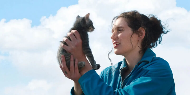 Eva Victor holding up a cat in front of a blue sky and white puffy clouds