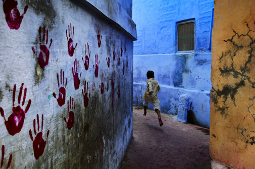a boy runs through an alley, the wall covered in painted handprints